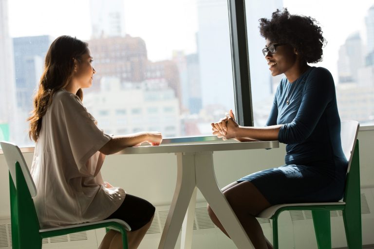 Two women in conversation across a table by a sunlit window